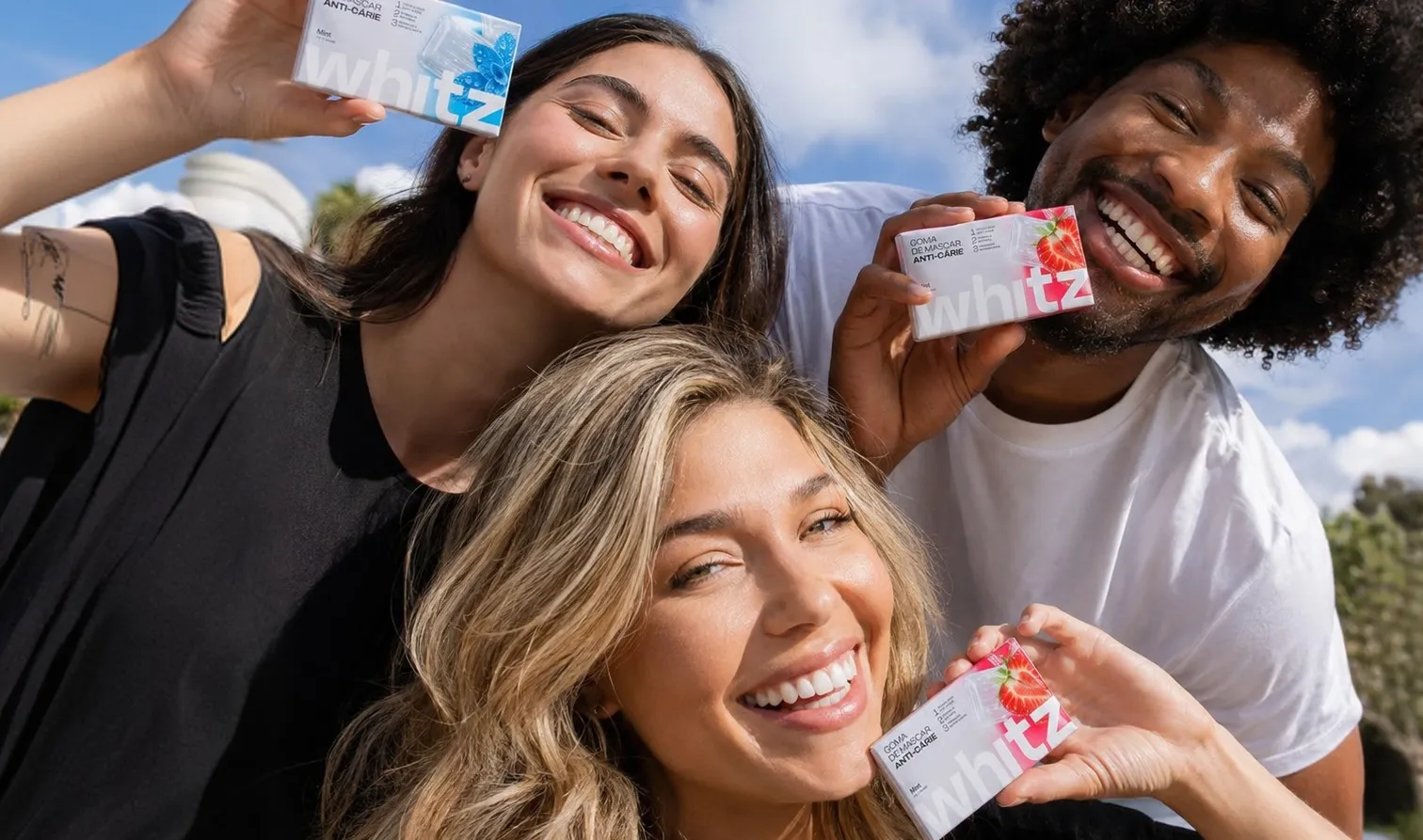 Três amigos sorrindo ao ar livre segurando embalagens de Whitz